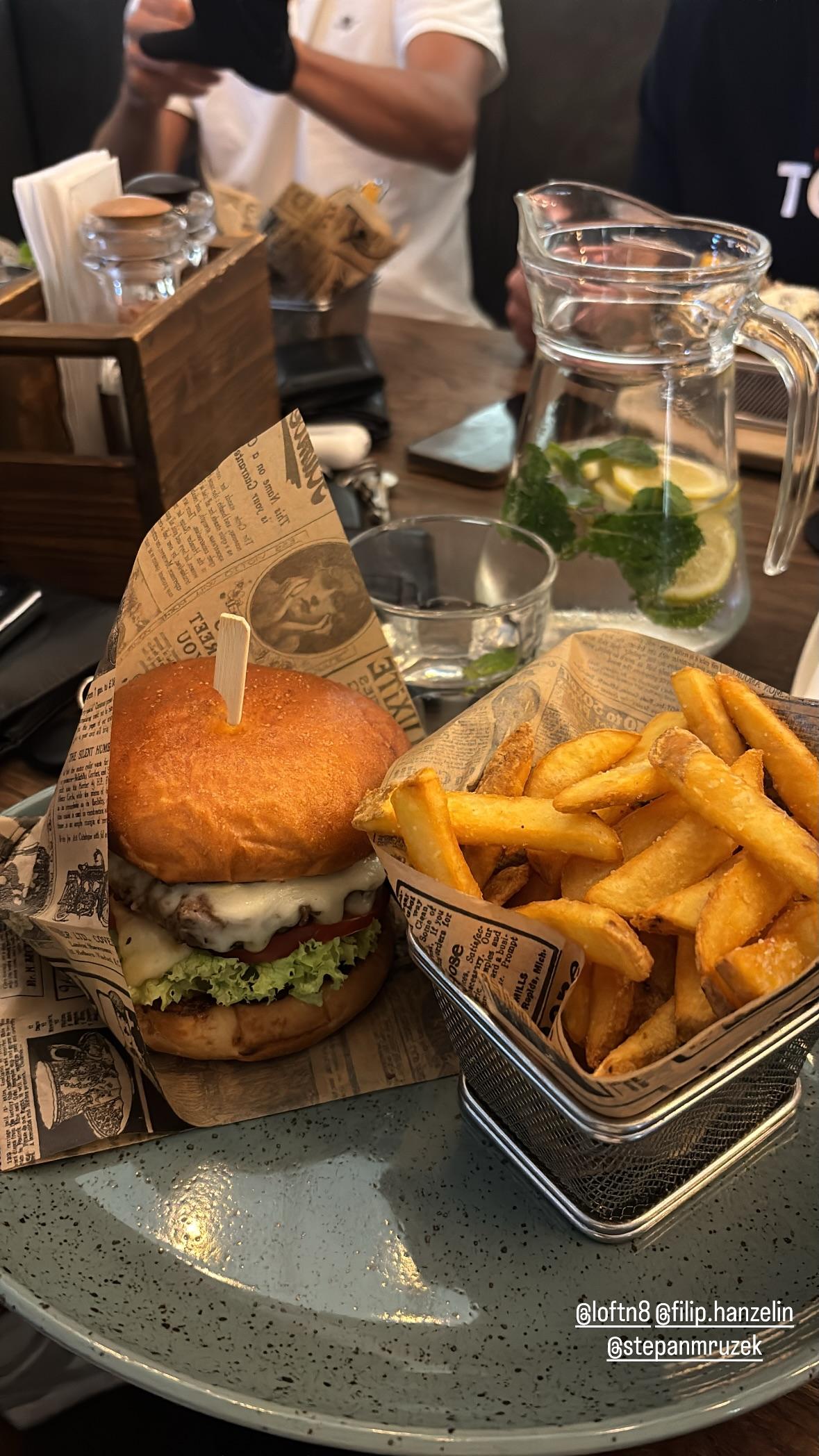Instagramové story z Loft N8: cheeseburger a košík hranolek na novinovém papíru, vedle džbán s vodou s citronem a mátou